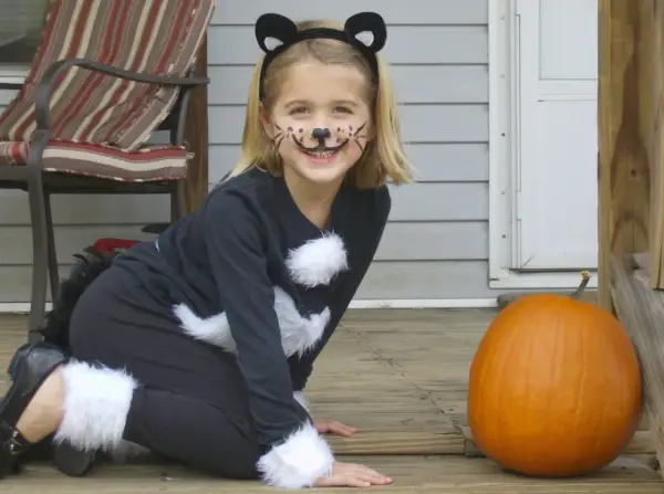 Bambina con trucco da gatto e orecchie, pronta per vestiti carnevale veloci fai da te bambini. Zucca arancione accanto.